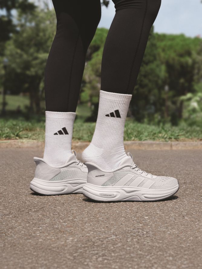The image shows a person's lower body wearing black leggings and white athletic shoes with the Adidas logo on a paved path surrounded by a lush, green environment.