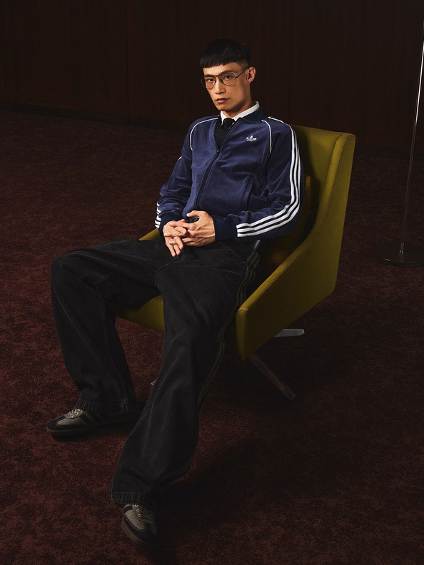 A person wearing a navy blue tracksuit jacket and black pants is seated in a yellow armchair against a dark background.