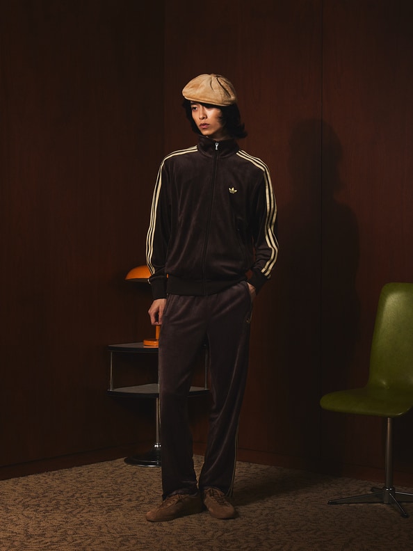 A person wearing a black tracksuit and a cap stands in a dimly lit room, with a chair and a green object visible in the background.