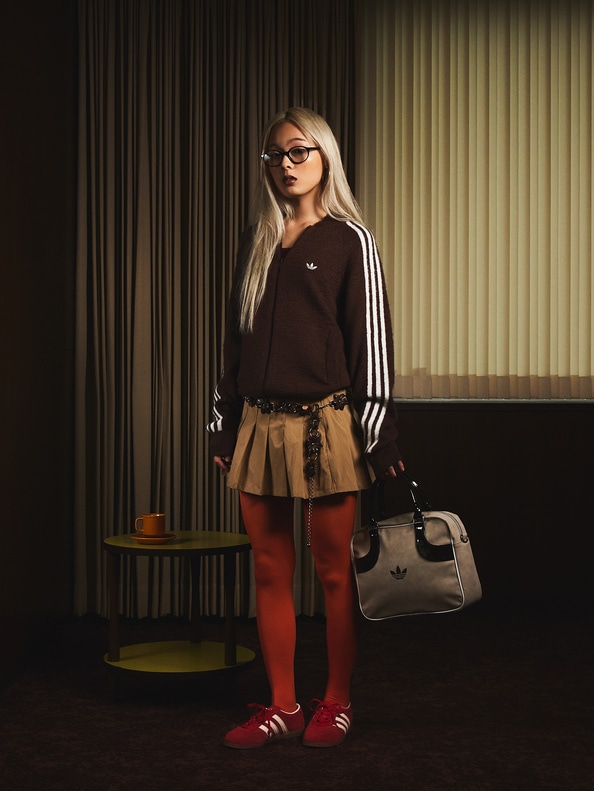 A young woman with long blonde hair stands in a dimly lit room, wearing a brown sweater, red pants, and red sneakers, holding a black handbag.