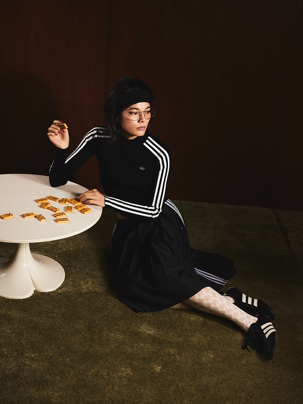 A young woman in a black dress and striped socks sits on the floor, surrounded by what appears to be cookies or pastries on a white pedestal table.