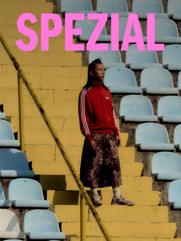 Una persona con una chaqueta roja y un pantalón corto estampado está de pie en unas escaleras frente a un gran cartel amarillo con la palabra "SPEZIAL".