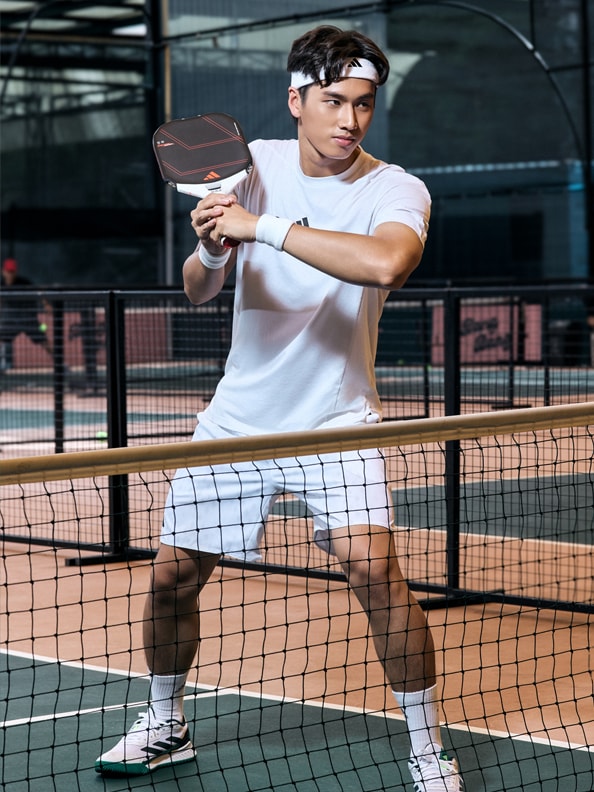 A young man in athletic attire stands on a tennis court, holding a tennis racket and preparing to serve the ball.
