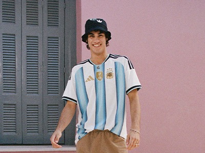 Un hombre joven con una camiseta de la selección argentina y una gorra de béisbol está de pie frente a una pared de color rosa con persianas.