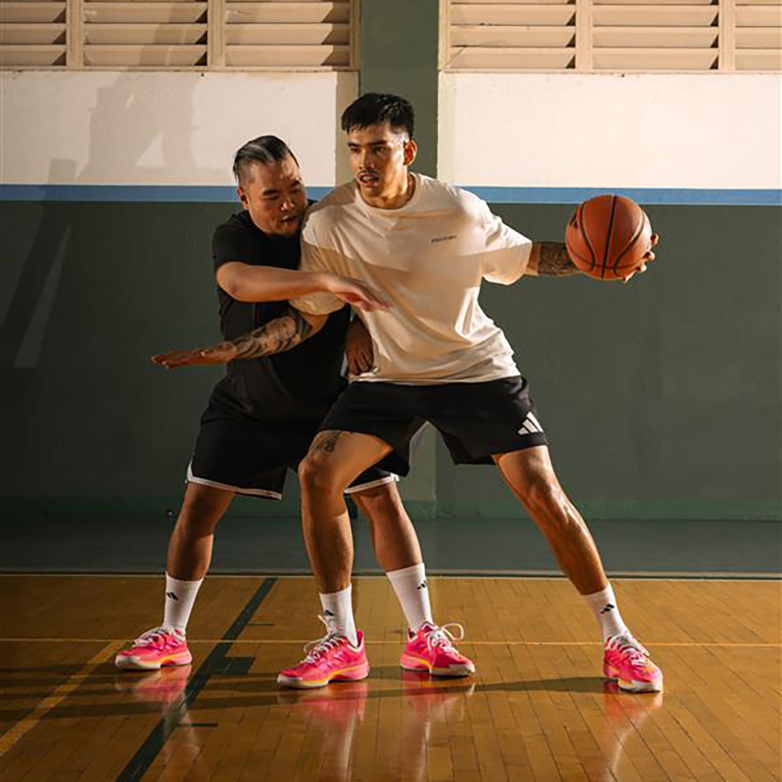 Two individuals, one in a white shirt and the other in a black shirt, are engaged in a basketball game on a wooden court with a basketball hoop visible in the background.