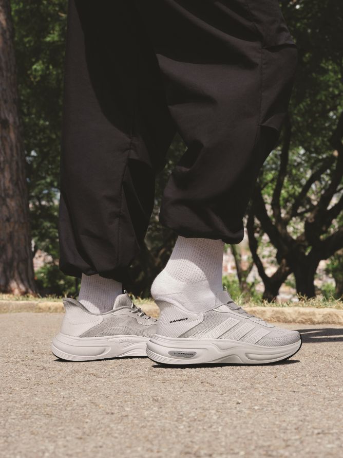 The image shows a person's lower body wearing black pants and gray sneakers standing on a paved path surrounded by trees and foliage.