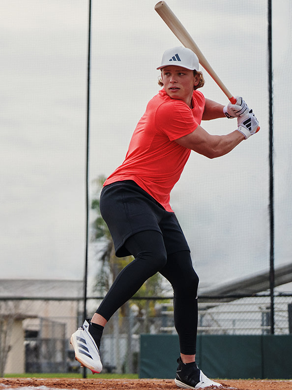 Una persona con una camicia rossa e un cappellino da baseball sta agitando una mazza da baseball su un campo da baseball con una staccionata e edifici visibili sullo sfondo.
