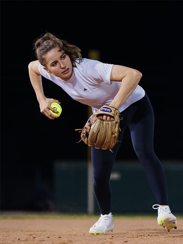 Una mujer atleta con una camiseta blanca y un pantalón negro, agachada en un campo de béisbol sujetando un guante y un balón.