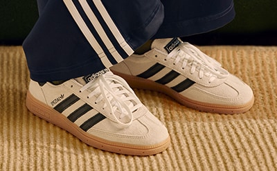 The image shows a pair of white and black Adidas sneakers with a gum sole, resting on a wooden floor.