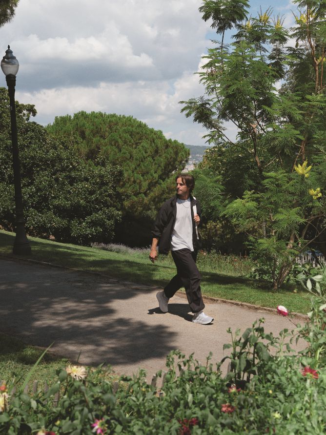 A person is walking on a paved path surrounded by lush greenery, including trees and shrubs, against a backdrop of a cloudy sky.
