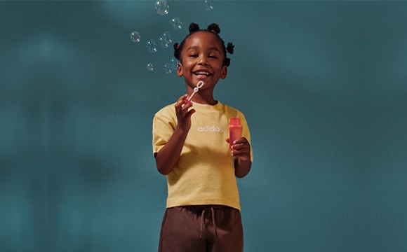 A young girl with braided hair wearing a yellow shirt is sticking out her tongue, surrounded by water droplets against a teal background.