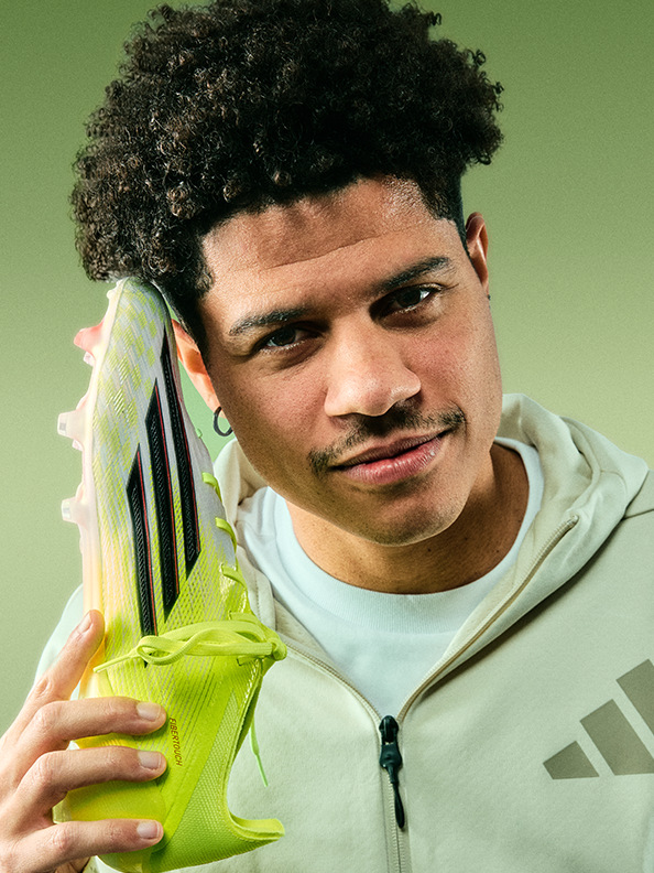 A young man with curly hair and a serious expression holds a green and white athletic shoe in front of a plain green background.
