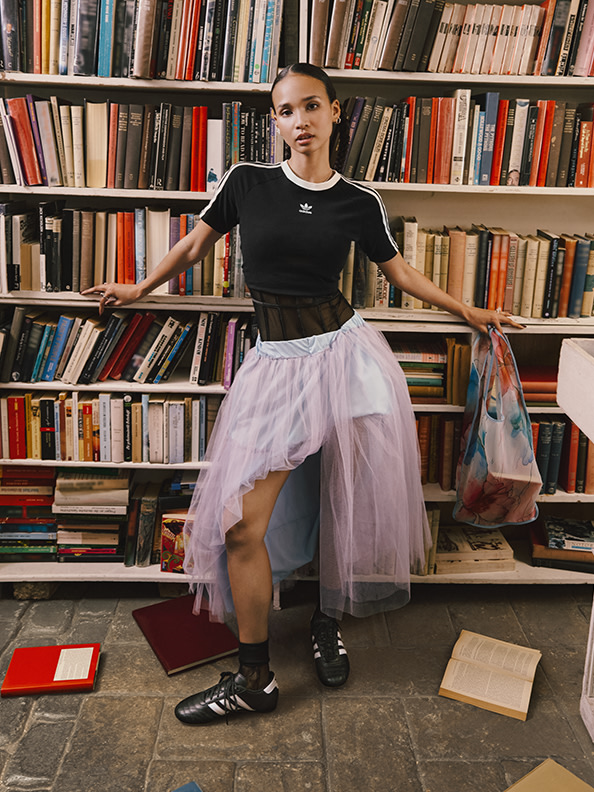 A young person wearing a black top and a large, puffy pink tulle skirt stands in front of a bookshelf filled with various books.