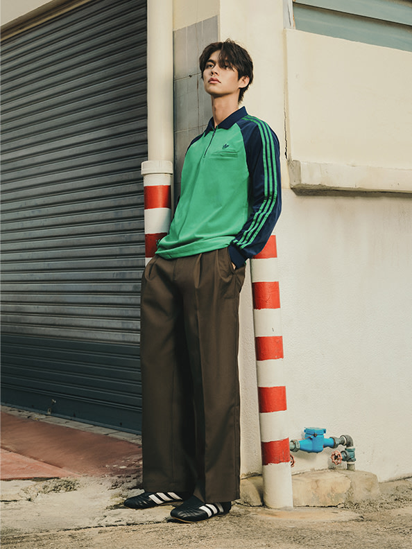 A young person wearing a green shirt and brown pants stands in front of a metal shutter, with red and white striped barriers visible in the background.