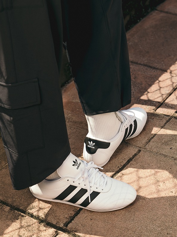 The image shows a pair of white and black Adidas sneakers on a person's feet, standing on a tiled floor.