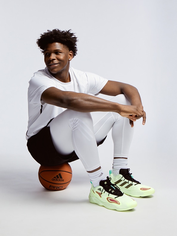 A young man with curly hair wearing a white shirt and black pants is sitting on a basketball while wearing bright green and black sneakers, against a plain white background.