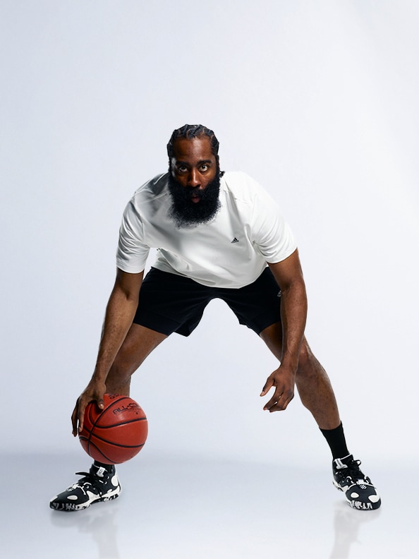 A man with a thick beard, wearing a white t-shirt and black shorts, is crouched down and holding a basketball in front of a plain white background.