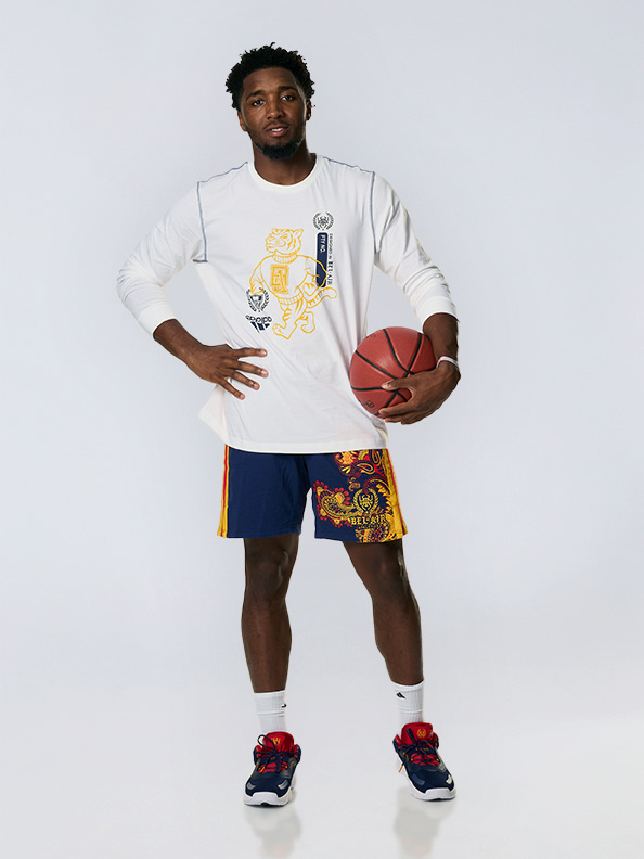 A young man wearing a white t-shirt with graphics, navy blue shorts, and red and blue sneakers, holding a basketball in a studio setting with a plain background.