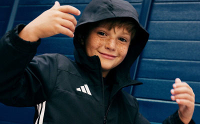 A young person wearing a black hooded jacket stands in front of a blue wall, making a thumbs-up gesture with both hands.