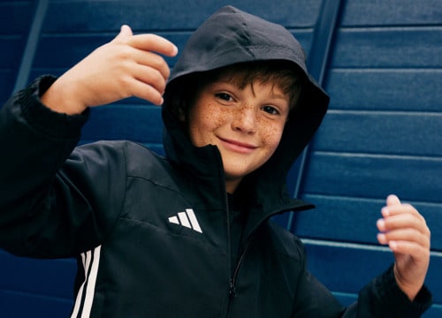 A young person wearing a black hooded jacket stands in front of a blue wall, making a gesture with their hands.