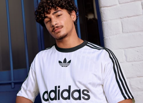 A young man with curly hair wearing a white Adidas shirt stands in front of a blue and white brick wall.