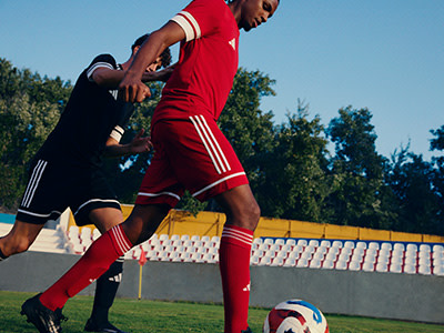 A person in a red sports uniform is dribbling a soccer ball on a sports field with a fence and trees in the background.