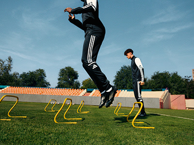 Two people in athletic attire are jumping over yellow hurdles on a grassy field, with a colorful background of buildings and trees.