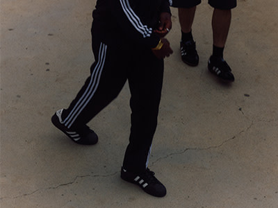 The image shows the lower body of two people wearing athletic clothing, including black pants with white stripes and black shoes, walking on a concrete surface.