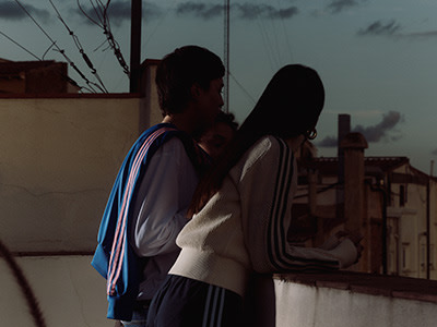 Two individuals, a man and a woman, stand together in a dimly lit setting, with a cityscape visible through a window in the background.