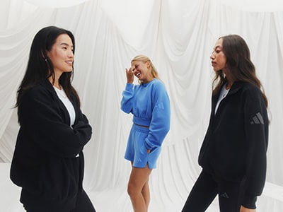 Three women in stylish outfits stand against a white backdrop, one wearing a bright blue dress and the others in black coats.