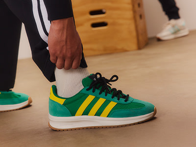 A pair of green and yellow Adidas sneakers on a wooden floor, with a person's legs and feet visible in the background.