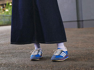 The image shows a person's lower body wearing a long, dark-colored skirt and a pair of blue and white sneakers standing on a paved surface with a blurred background.