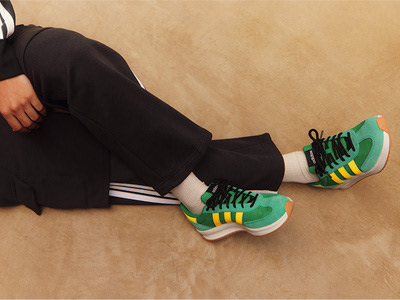 The image shows a person's legs and feet wearing black pants and green and yellow sneakers, sitting on a beige floor.