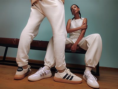 The image depicts a woman wearing a white outfit, including a sleeveless top and loose-fitting pants, sitting on a bench against a teal-colored wall. Her feet are visible, wearing white sneakers with black stripes.