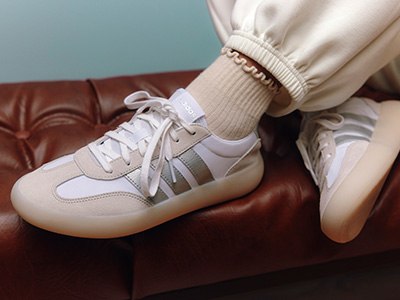 The image shows a pair of white and gray sneakers on a leather couch, with a person's legs and socks visible in the foreground.