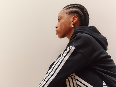 A person with braided hair wearing a black hoodie stands against a plain background.