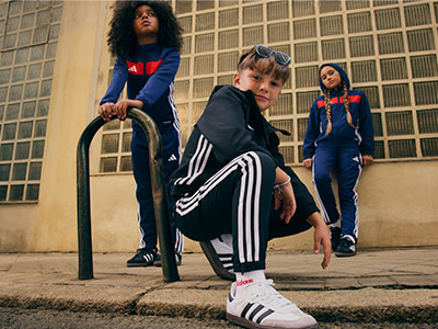 Three individuals, two women and one man, are standing in front of a building with a grid-like pattern on the walls. The individuals are wearing athletic clothing, including tracksuits and sneakers, and appear to be in a casual, relaxed pose.