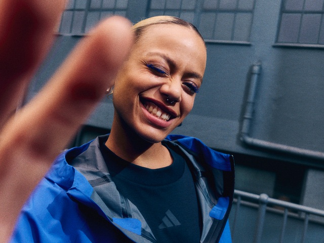 A smiling woman in a blue jacket stands in front of a gray building.