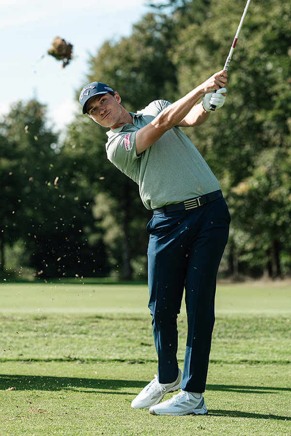 A person in a golf outfit is swinging a golf club on a grassy golf course surrounded by trees.