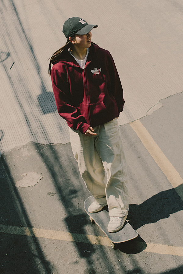 A person wearing a red jacket and a baseball cap stands in front of a concrete wall, casting a long shadow on the ground.
