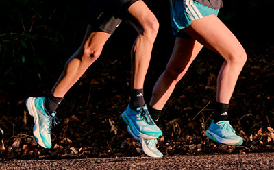 The image shows a pair of legs wearing colorful athletic shoes running on a dirt path surrounded by foliage.