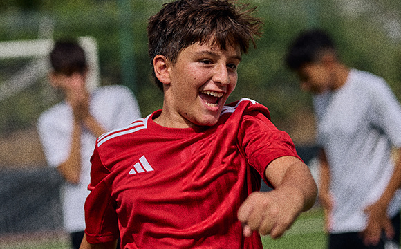 A young male athlete in a red sports jersey is enthusiastically celebrating on a grassy field, with other players visible in the background.