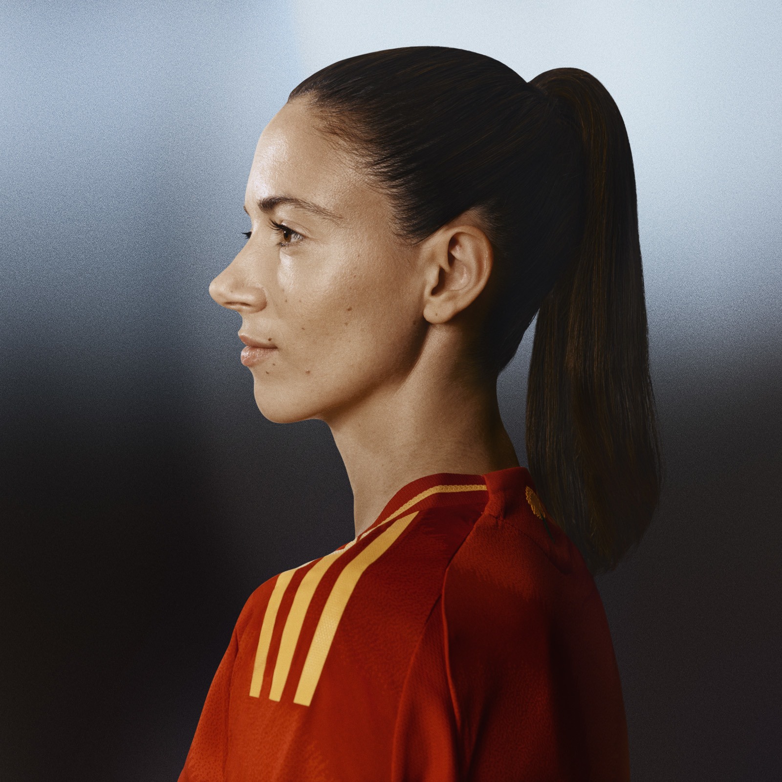 En person som bär en röd och gul sporttröja står i profil mot en mörk bakgrund, och deras uttryck är seriöst och fokuserat.