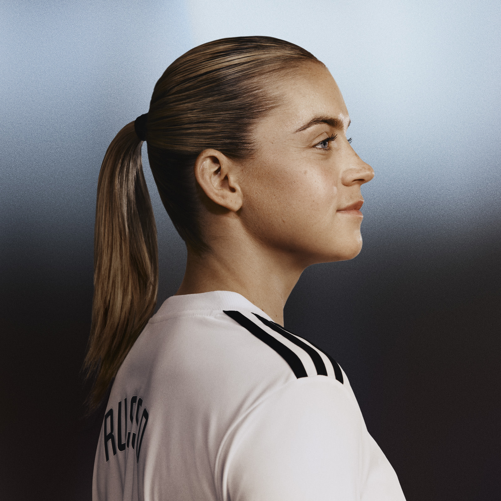 En ung kvinna med långt, mörkt hår i en hästsvans, iklädd en vit sporttröja, står i profil mot en mörk bakgrund.