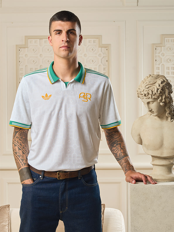 Un ragazzo con i tatuaggi sulle braccia si trova di fronte a un muro decorativo, indossa una polo bianca con dettagli verdi e gialli.