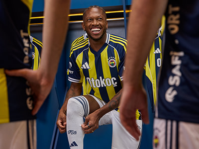 A smiling man wearing a yellow and blue soccer jersey stands in a locker room surrounded by sports equipment and team gear.
