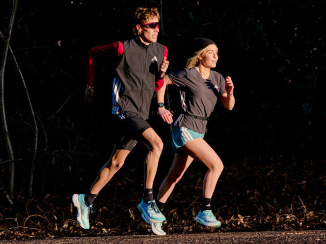 Two people, a man and a woman, are running together in a dark outdoor setting, with trees and foliage visible in the background.