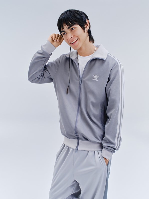 A young person wearing a gray Adidas tracksuit stands against a plain white background, smiling and gesturing with their hand.