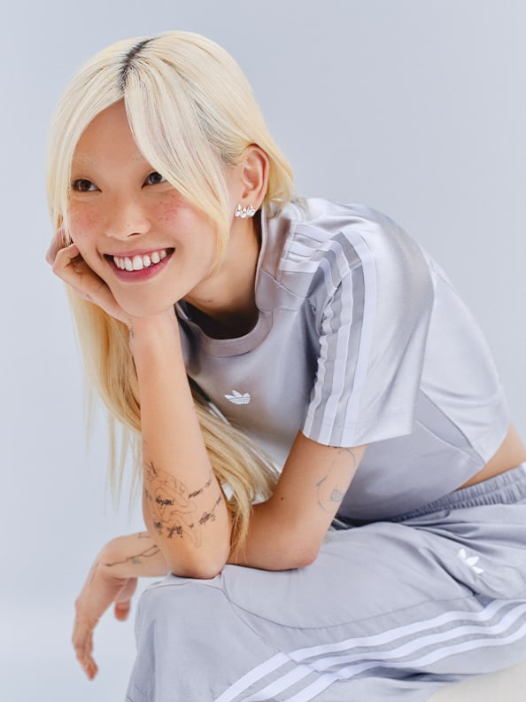 A smiling young woman with blonde hair wearing a white and gray athletic outfit sits on the floor against a plain gray background.