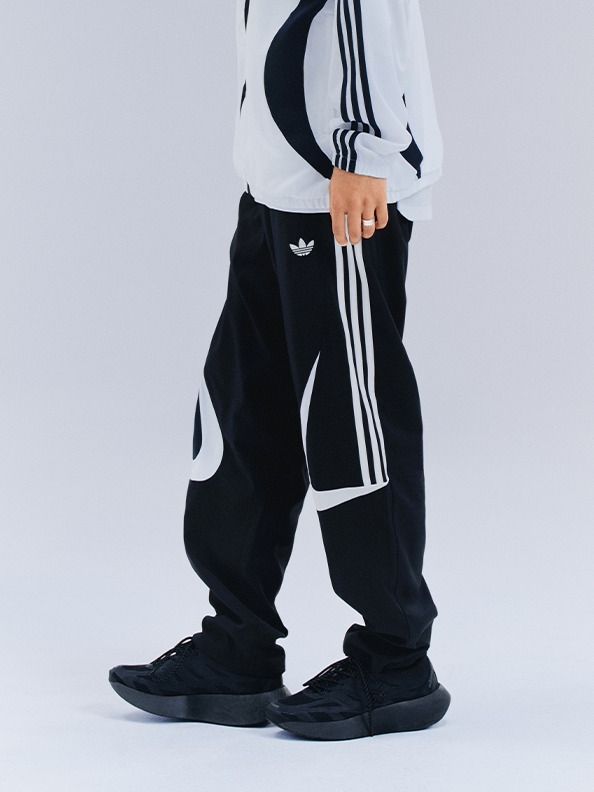 The image shows a person wearing a white and black patterned top and black pants with the Adidas logo, standing against a plain white background.